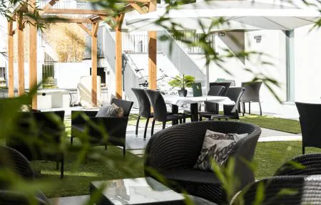 A patio designed as a restaurant terrace, with chairs and tables, a wicker lounge area, white shade sails providing sun protection, tiled flooring, and green spaces.