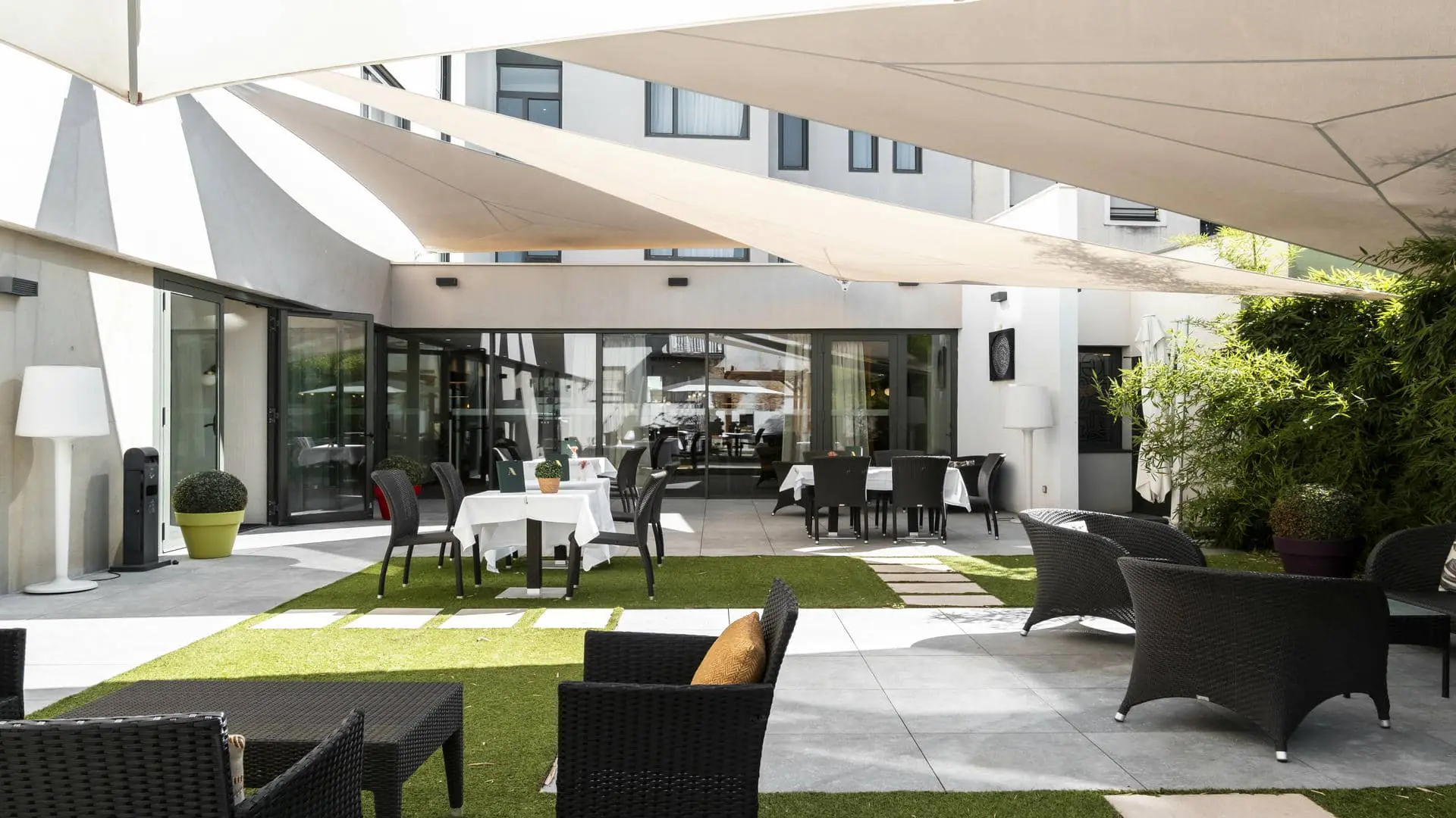 Un patio aménagé en terrasse de restaurant avec des chaises et des tables, un coin salon en osier, protégé du soleil par des toiles tendues blanches, un sol carrelé et des espaces verts.