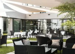 A patio designed as a restaurant terrace, with chairs and tables, a wicker lounge area, white shade sails providing sun protection, tiled flooring, and green spaces.