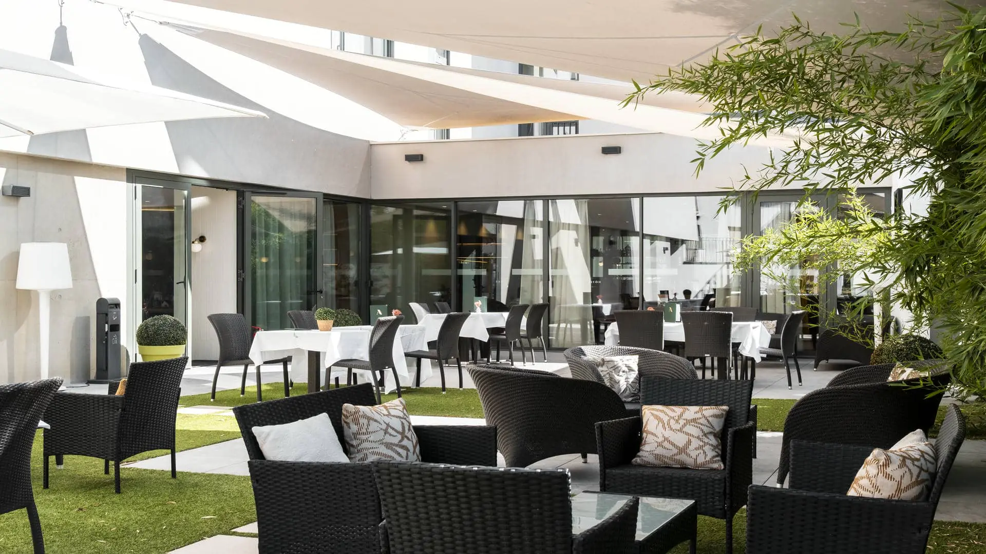 A patio designed as a restaurant terrace, with chairs and tables, a wicker lounge area, white shade sails providing sun protection, tiled flooring, and green spaces.