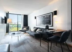 A hotel suite with a lounge area, a desk, and a stylish wall-mounted television, filled with natural light from a large floor-to-ceiling window overlooking the trees.