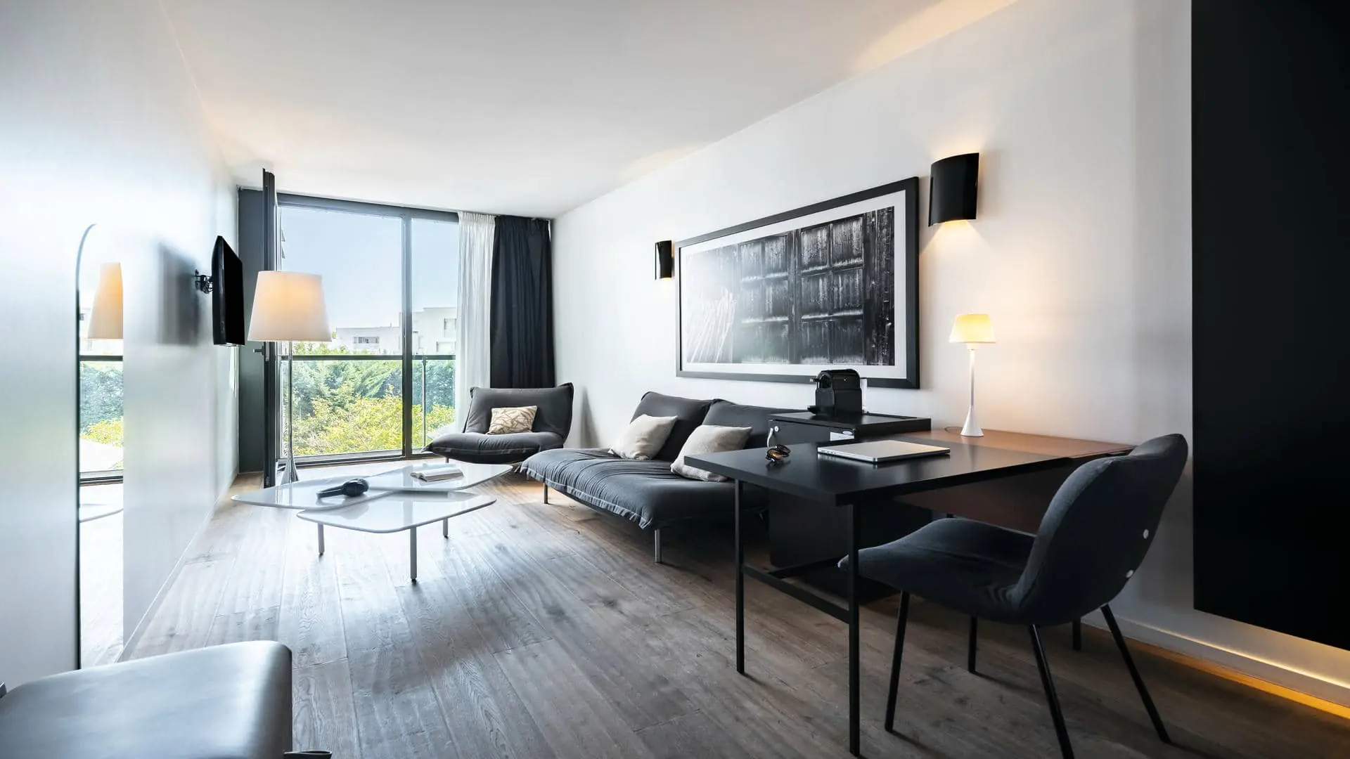 A hotel suite with a lounge area, a desk, and a stylish wall-mounted television, filled with natural light from a large floor-to-ceiling window overlooking the trees.