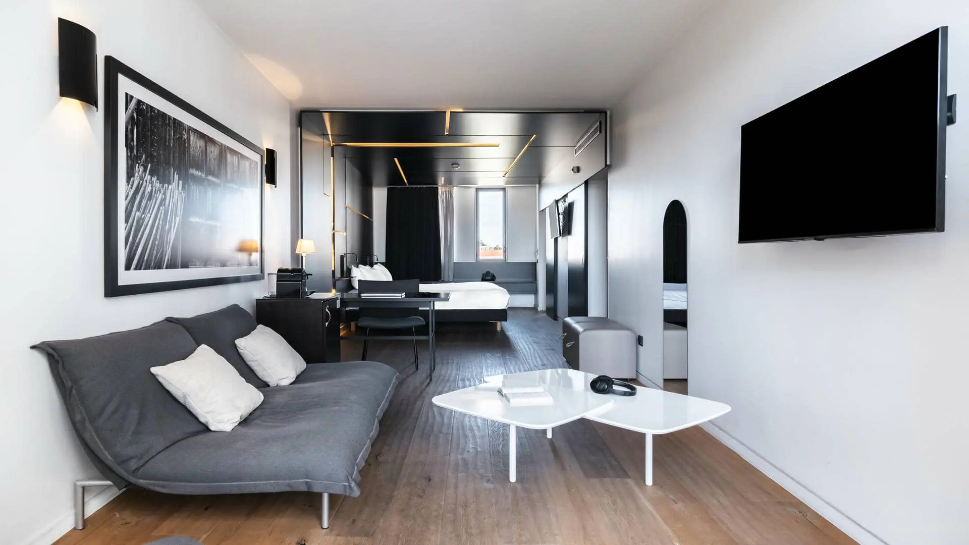 A hotel suite with a lounge area, a desk, and a stylish wall-mounted television, filled with natural light from a large floor-to-ceiling window overlooking the trees.