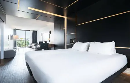 A hotel suite with a lounge area, a desk, and a stylish wall-mounted television, filled with natural light from a large floor-to-ceiling window overlooking the trees.