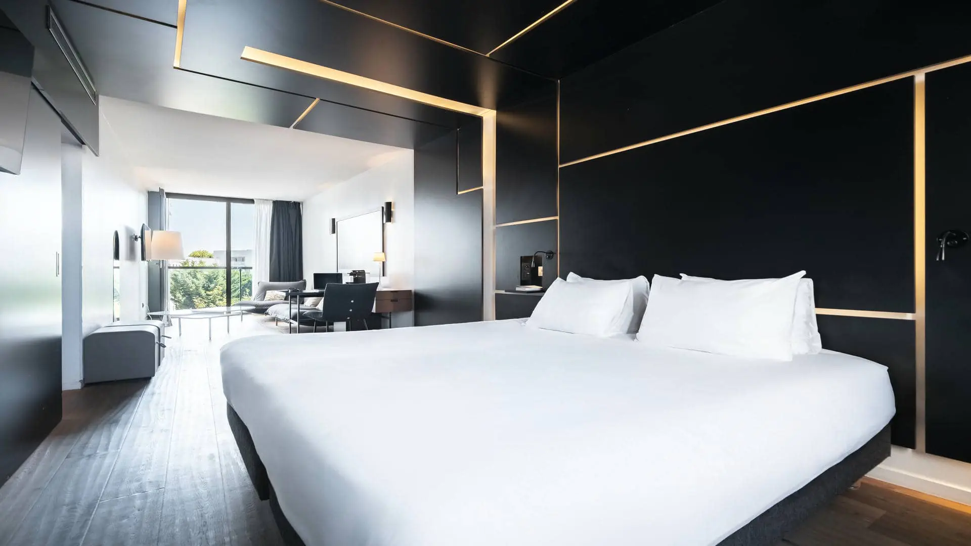 A hotel suite with a lounge area, a desk, and a stylish wall-mounted television, filled with natural light from a large floor-to-ceiling window overlooking the trees.