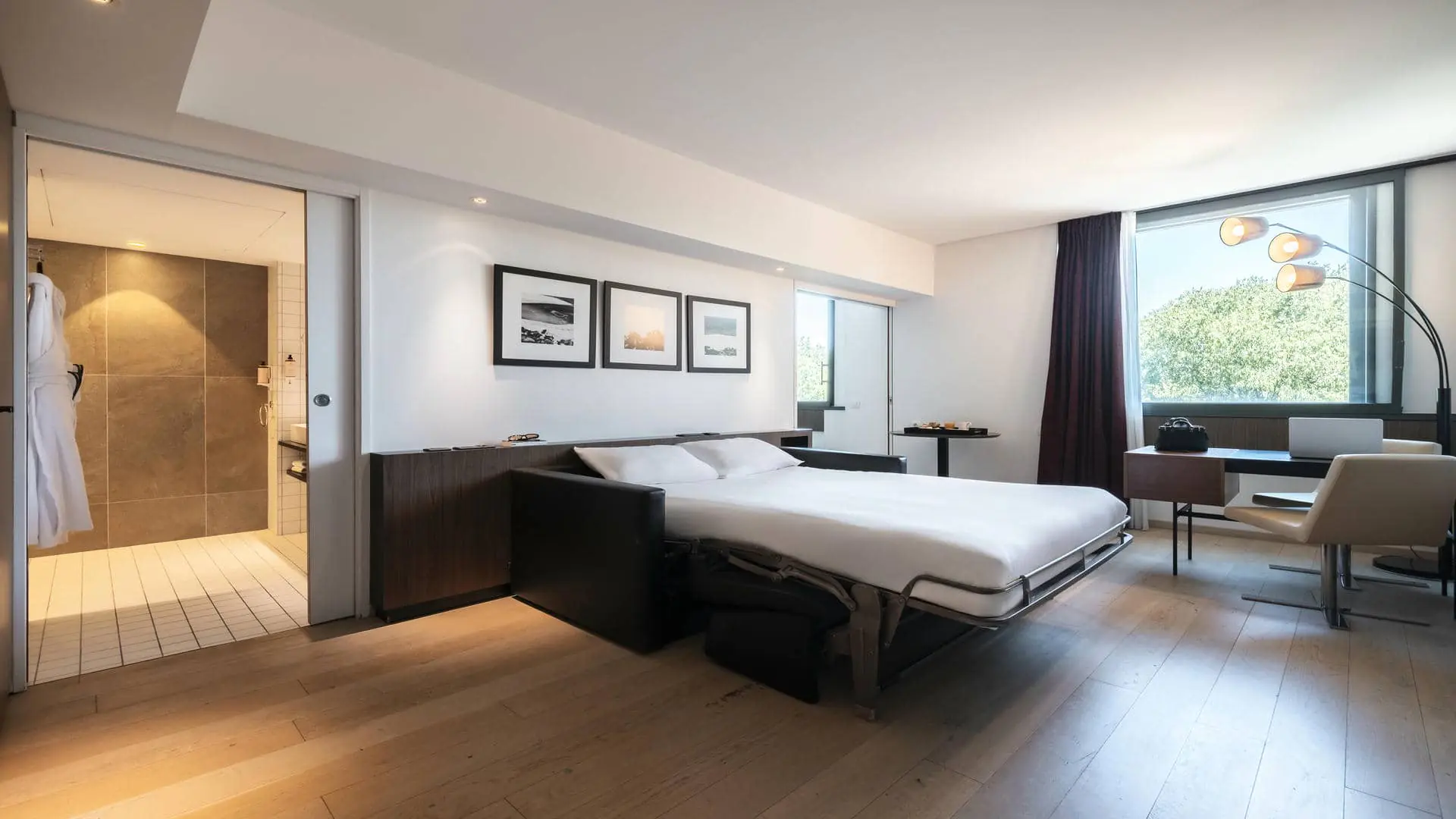 View of the hotel suite featuring a sofa bed prepared for sleeping, a desk positioned by the window overlooking the greenery, and a bathroom equipped with bathrobes.