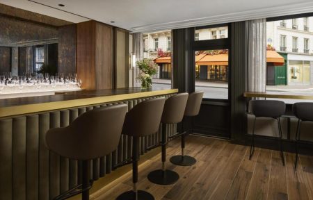 Élégant bar d’hôtel avec long comptoir en bois sombre et laiton, fauteuils hauts taupe, éclairage chaleureux, verrerie, compositions florales et grandes baies vitrées donnant sur une rue parisienne.