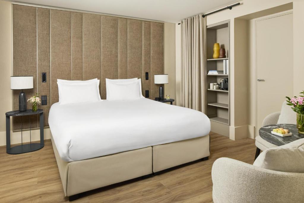 Chambre d’hôtel de luxe contemporaine avec lit king-size, tête de lit beige texturée, tables de chevet sombres, parquet chaleureux, rideaux aux tons naturels et petit espace salon avec table en marbre, mignardises et fleurs fraîches.
