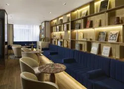 Élégant lounge d’hôtel avec banquette en velours bleu profond, bibliothèque en bois éclairée, fauteuils beige, tables rondes en marbre, compositions florales et douce lumière naturelle.