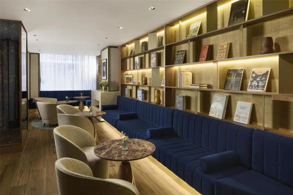 Élégant lounge d’hôtel avec banquette en velours bleu profond, bibliothèque en bois éclairée, fauteuils beige, tables rondes en marbre, compositions florales et douce lumière naturelle.