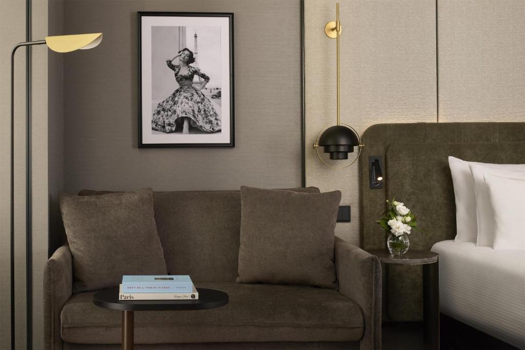 Bedroom with a king-size bed dressed in white linens and an olive green upholstered headboard. A two-seater taupe velvet sofa with matching cushions and a round coffee table sits beside the bed. Textured grey-beige paneled walls, black and gold wall lights, a small bedside table with a flower vase, and a framed photograph above the sofa complete the room.