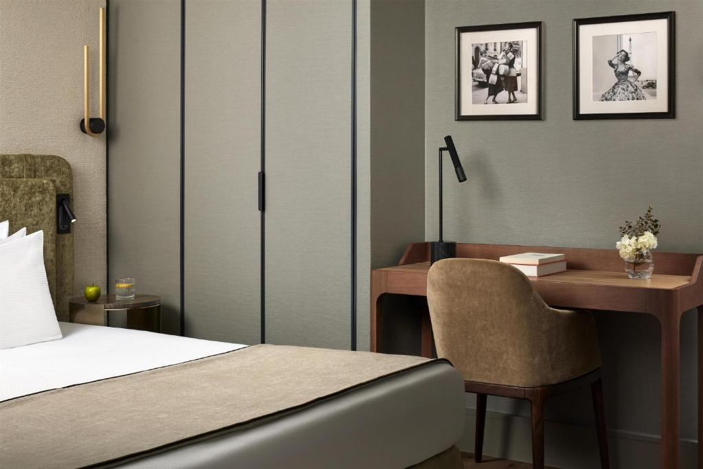 Elegant contemporary guest room featuring a double bed dressed in crisp white linens with a soft beige throw. A refined wooden writing desk, paired with a velvet armchair and reading lamp, creates a warm work area. Black-and-white photographs add an artistic, Parisian touch.