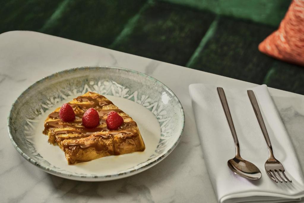 Dessert served on a delicately patterned ceramic plate: (french toast) pastry drizzled with caramel sauce and topped with three raspberries. The plate is set on a white marble table, with metal cutlery placed on a white napkin.