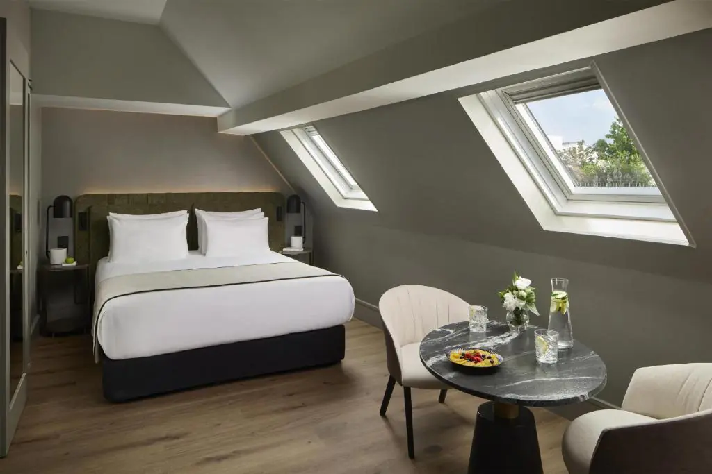 Elegant, light-filled attic room featuring a refined double bed with a padded headboard and crisp white linens. A cozy seating area with a marble table, comfortable armchairs, chilled water and fresh fruit creates an intimate and sophisticated hotel atmosphere.