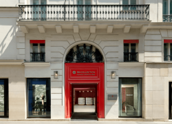 Elegant Parisian hotel façade bathed in natural daylight, featuring a refined white stone exterior with classic architectural detailing, wrought-iron balconies and tall French windows accented with deep red trim. A striking red entrance framed by ornate stonework and a decorative wrought-iron arch creates a sophisticated focal point, while large ground-floor windows reveal glimpses of the luxurious interior. The harmonious blend of traditional Parisian architecture and contemporary hospitality design evokes the refined atmosphere of a prestigious boutique hotel.