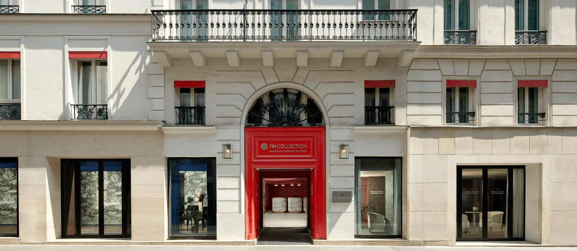 Elegant Parisian hotel façade bathed in natural daylight, featuring a refined white stone exterior with classic architectural detailing, wrought-iron balconies and tall French windows accented with deep red trim. A striking red entrance framed by ornate stonework and a decorative wrought-iron arch creates a sophisticated focal point, while large ground-floor windows reveal glimpses of the luxurious interior. The harmonious blend of traditional Parisian architecture and contemporary hospitality design evokes the refined atmosphere of a prestigious boutique hotel.