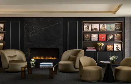 Elegant and sophisticated hotel lounge featuring a dramatic black marble fireplace, surrounded by refined built-in dark wood bookshelves filled with art, fashion and design books. Several curved taupe velvet armchairs are arranged around low marble and dark wood tables, adorned with delicate floral arrangements and carefully selected coffee-table books. Warm recessed lighting, golden accents and the glowing fireplace create an intimate, luxurious and contemporary atmosphere.