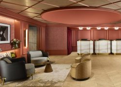 Elegant and spacious luxury hotel lobby in warm terracotta and burgundy tones, featuring refined wood-panelled walls, softly illuminated recessed shelves and sophisticated contemporary lighting. A stylish reception area with three curved marble and brass-fronted desks occupies the background, while a selection of black leather and patterned armchairs, rounded lounge chairs and small sculptural tables create inviting seating areas in the foreground. A large decorative circular ceiling feature, soft ambient lighting, framed fashion artwork and fresh floral arrangements enhance the chic Parisian atmosphere.