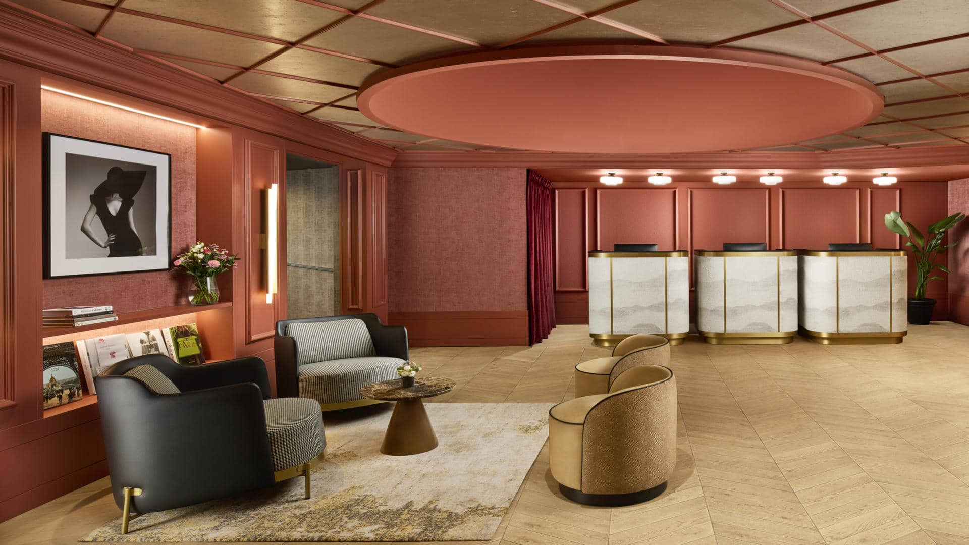 Elegant and spacious luxury hotel lobby in warm terracotta and burgundy tones, featuring refined wood-panelled walls, softly illuminated recessed shelves and sophisticated contemporary lighting. A stylish reception area with three curved marble and brass-fronted desks occupies the background, while a selection of black leather and patterned armchairs, rounded lounge chairs and small sculptural tables create inviting seating areas in the foreground. A large decorative circular ceiling feature, soft ambient lighting, framed fashion artwork and fresh floral arrangements enhance the chic Parisian atmosphere.