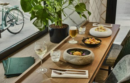 Wooden restaurant table with green and grey fabric armchairs, set beside a large window. The table is set with glasses, gold-colored cutlery, white napkins and several dishes, accompanied by a glass of beer and a glass of white wine. Large green plant and bicycle visible outside.