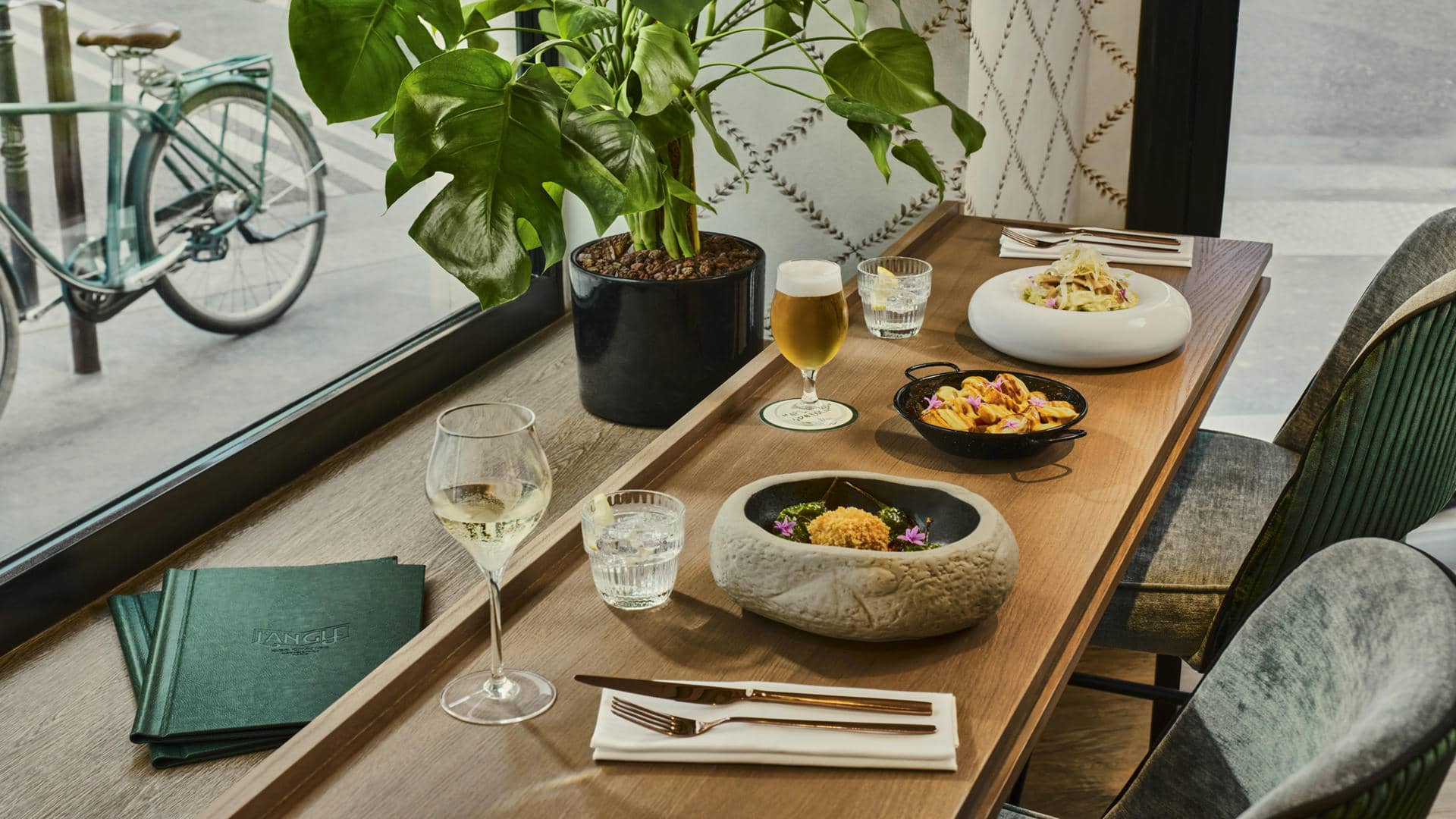 Wooden restaurant table with green and grey fabric armchairs, set beside a large window. The table is set with glasses, gold-colored cutlery, white napkins and several dishes, accompanied by a glass of beer and a glass of white wine. Large green plant and bicycle visible outside.