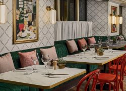Chic retro-style restaurant with dark green velvet banquettes and blush pink and terracotta cushions. White marble tables with gold trim, red chairs, white diamond-patterned wallpaper and glass and brass wall lights. Tables set with glasses, cutlery and small decorative plants.