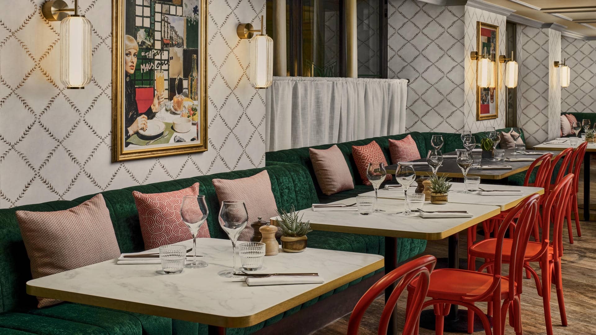 Chic retro-style restaurant with dark green velvet banquettes and blush pink and terracotta cushions. White marble tables with gold trim, red chairs, white diamond-patterned wallpaper and glass and brass wall lights. Tables set with glasses, cutlery and small decorative plants.