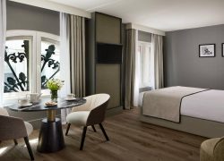 Room with a large double bed, light grey headboard and white bedding with a beige bedspread. Taupe-grey walls, wooden flooring and beige blackout curtains around two windows with black wrought-iron railings. Seating area with two cream armchairs and a round black marble table with a gold and black base, facing a wall-mounted TV framed in dark wood.