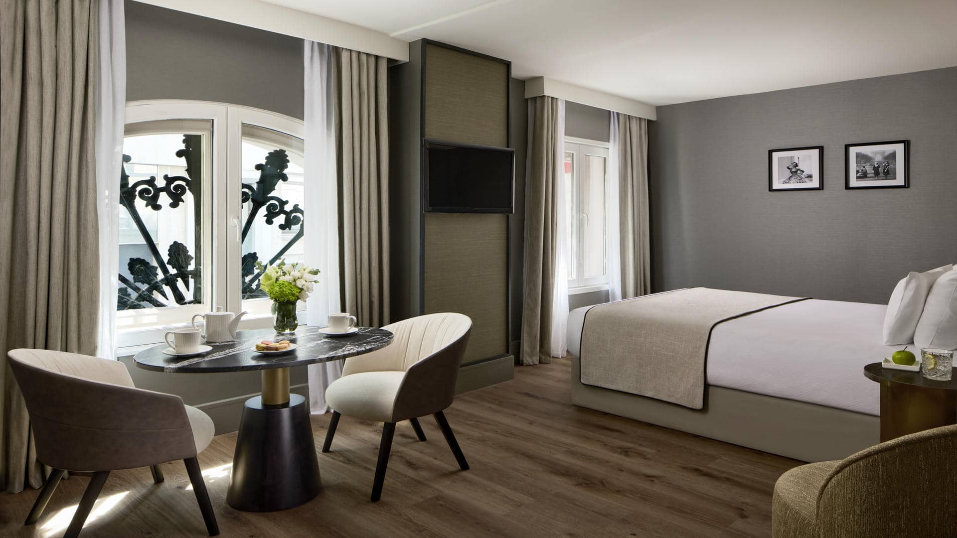 Room with a large double bed, light grey headboard and white bedding with a beige bedspread. Taupe-grey walls, wooden flooring and beige blackout curtains around two windows with black wrought-iron railings. Seating area with two cream armchairs and a round black marble table with a gold and black base, facing a wall-mounted TV framed in dark wood.
