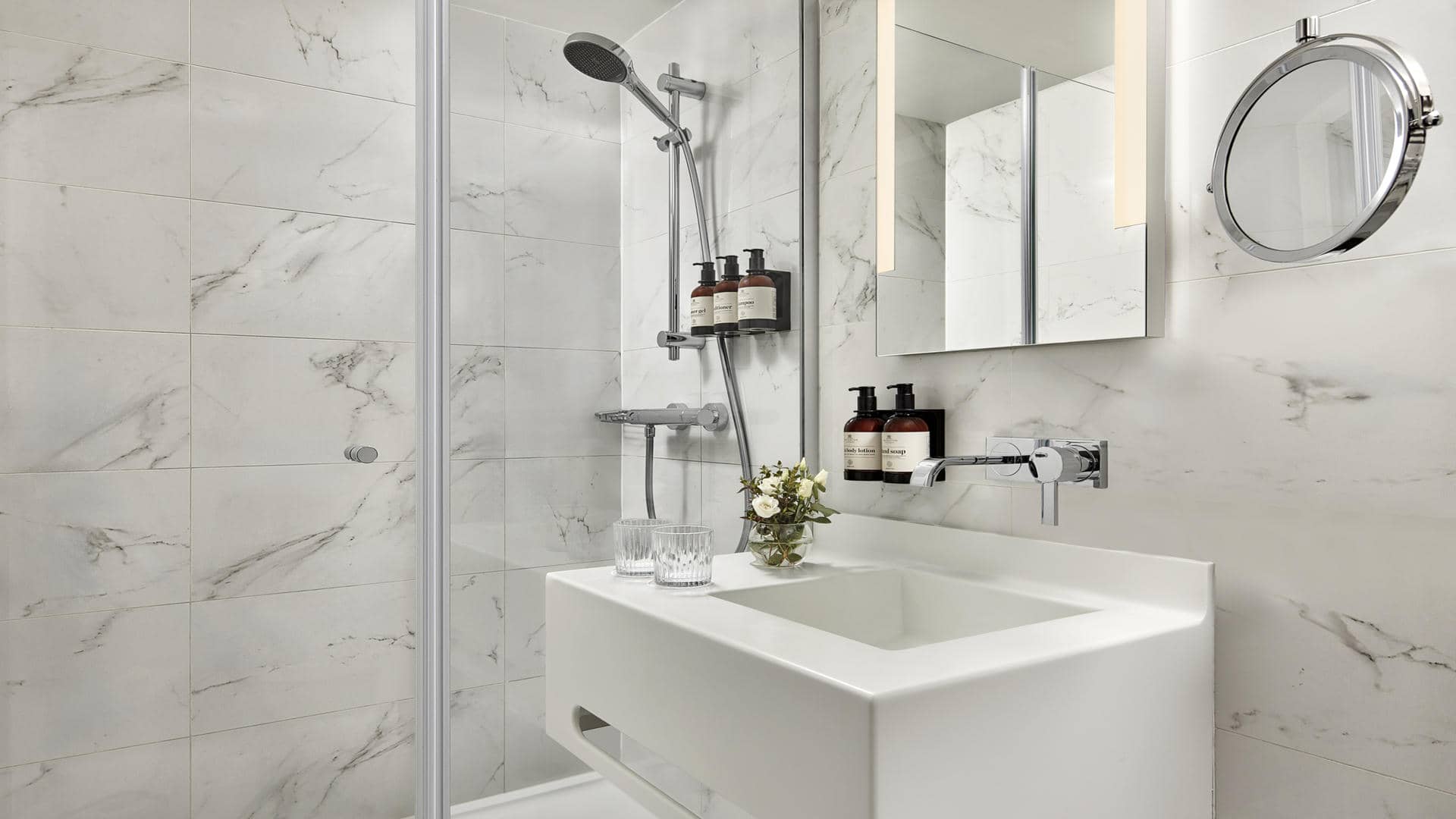 White-toned bathroom with white marble-effect wall and floor tiles featuring grey veining. Large rectangular white sink, wall-mounted mirror and round magnifying mirror, glass-enclosed shower with chrome fixtures and amber toiletry bottles.