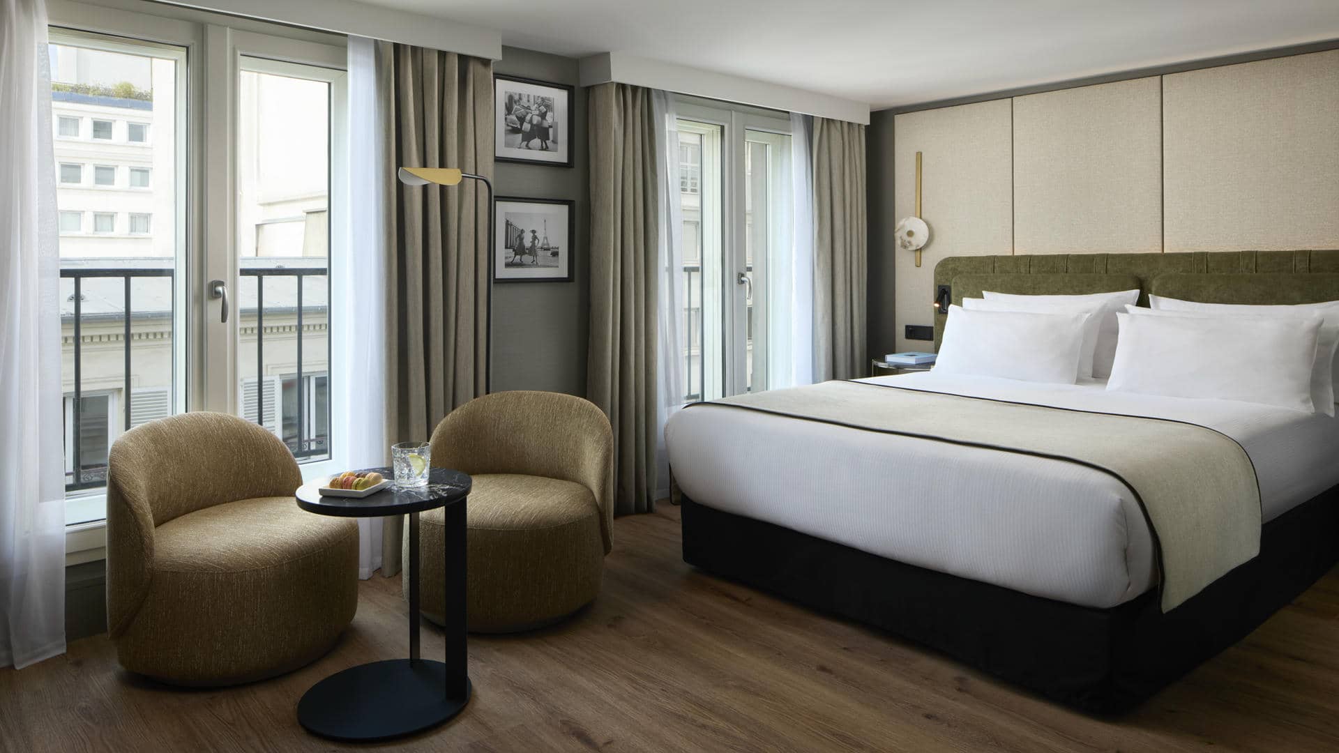 Room with a king-size bed, khaki green fabric headboard and white bedding. Two rounded beige fabric armchairs surround a small black round table with a plate and two glasses. Wooden flooring, grey and beige walls, taupe curtains and large windows overlooking the surrounding buildings.