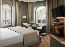 Room with a large double bed dressed in white and beige, wooden flooring and taupe-grey walls. Two arched windows with white shutters and views of Parisian façades, framed by beige blackout curtains. Seating area with two cream armchairs, a round black marble table and a black-and-white rug, facing a wall-mounted TV and a dark wood minibar cabinet.