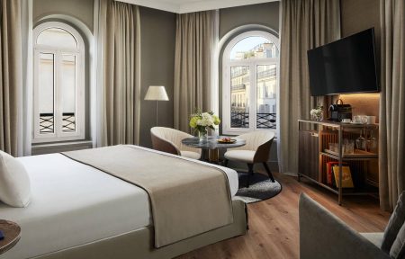 Room with a large double bed dressed in white and beige, wooden flooring and taupe-grey walls. Two arched windows with white shutters and views of Parisian façades, framed by beige blackout curtains. Seating area with two cream armchairs, a round black marble table and a black-and-white rug, facing a wall-mounted TV and a dark wood minibar cabinet.