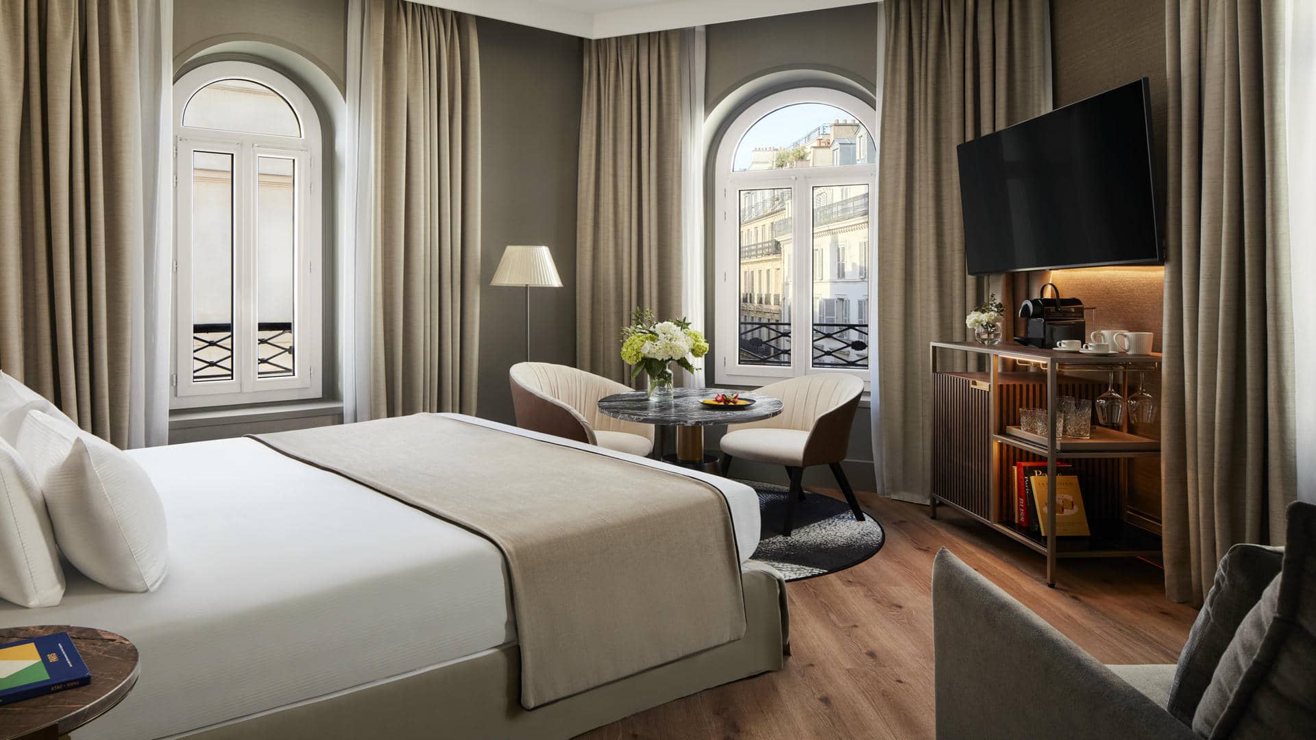 Room with a large double bed dressed in white and beige, wooden flooring and taupe-grey walls. Two arched windows with white shutters and views of Parisian façades, framed by beige blackout curtains. Seating area with two cream armchairs, a round black marble table and a black-and-white rug, facing a wall-mounted TV and a dark wood minibar cabinet.