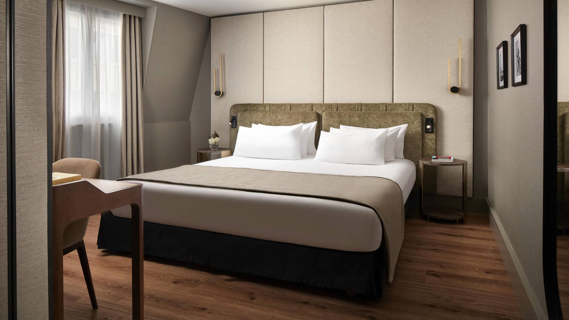 Room with a large double bed and a khaki-green fabric headboard, set against cream wall panels. White and beige bedding, wooden flooring, taupe-grey walls, beige blackout curtains and a matching armchair. Wooden desk, bedside tables and black and gold wall lights.