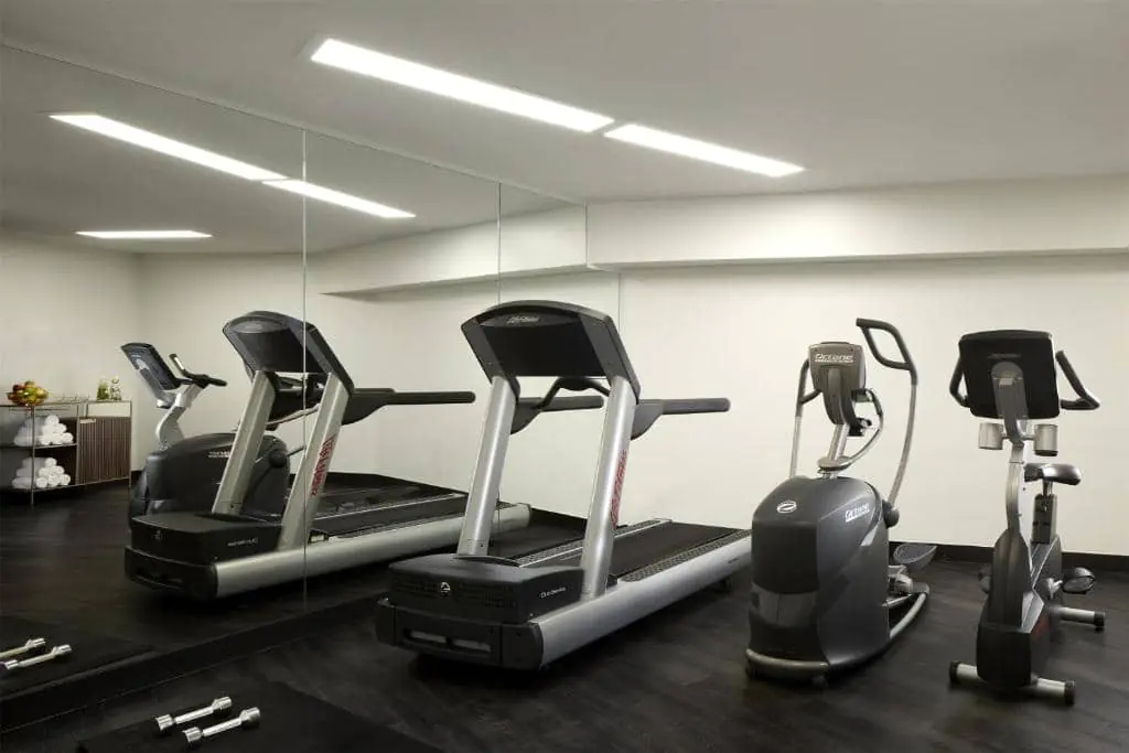 Modern fitness room with treadmills and elliptical machines on a black floor. Large wall mirror, white walls and ceiling spotlights. Dumbbells are placed on the floor.