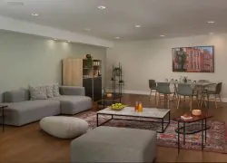Salon moderne comprenant un grand canapé gris rembourré avec des coussins géométriques, un pouf beige et un ottoman assorti. Une table basse en marbre avec une structure en métal noir est installée sur un tapis rouge à motifs, décorée de pommes vertes et de jus d’orange. De petites tables d’appoint noires accueillent des bougies et des fleurs fraîches. À l’arrière-plan, un meuble en bois aux étagères ouvertes, des plantes vertes et une table à manger entourée de chaises grises complètent l’espace. Un tableau encadré coloré, un mur vert sauge, un papier peint à motifs et un parquet chaleureux créent une atmosphère élégante et contemporaine.