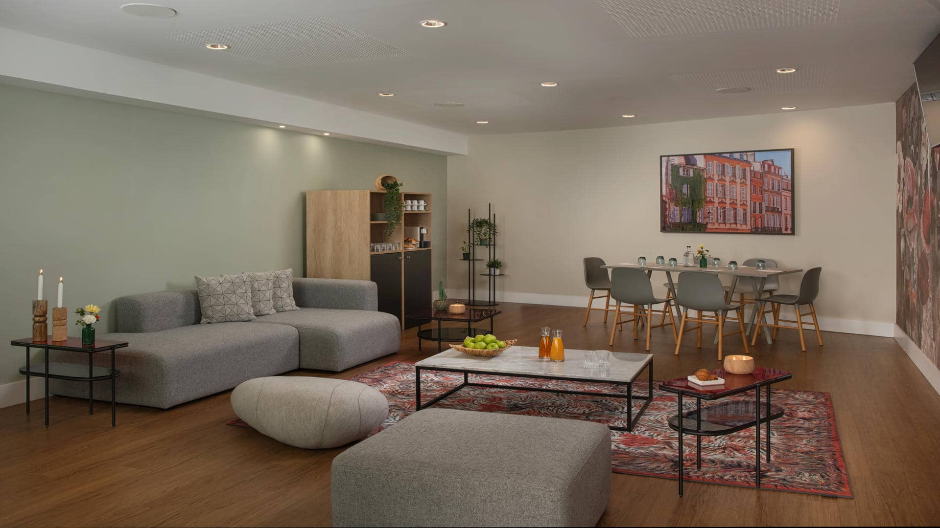 Salon moderne comprenant un grand canapé gris rembourré avec des coussins géométriques, un pouf beige et un ottoman assorti. Une table basse en marbre avec une structure en métal noir est installée sur un tapis rouge à motifs, décorée de pommes vertes et de jus d’orange. De petites tables d’appoint noires accueillent des bougies et des fleurs fraîches. À l’arrière-plan, un meuble en bois aux étagères ouvertes, des plantes vertes et une table à manger entourée de chaises grises complètent l’espace. Un tableau encadré coloré, un mur vert sauge, un papier peint à motifs et un parquet chaleureux créent une atmosphère élégante et contemporaine.