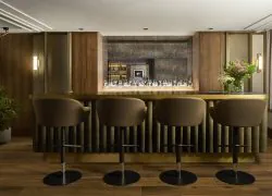 Élégant bar d’hôtel avec comptoir en bois sombre et laiton, quatre fauteuils hauts en velours brun, étagères éclairées, verrerie, compositions florales et grande fenêtre donnant sur une rue parisienne.
