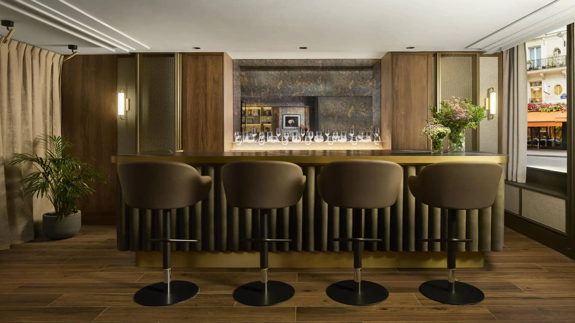 Élégant bar d’hôtel avec comptoir en bois sombre et laiton, quatre fauteuils hauts en velours brun, étagères éclairées, verrerie, compositions florales et grande fenêtre donnant sur une rue parisienne.