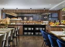 Restaurant d’hôtel pour groupes avec buffet en bois et pierre sombre proposant pains, viennoiseries, céréales, yaourts, fromages, charcuteries, fruits frais et jus, avec tables claires et assises en velours bleu.