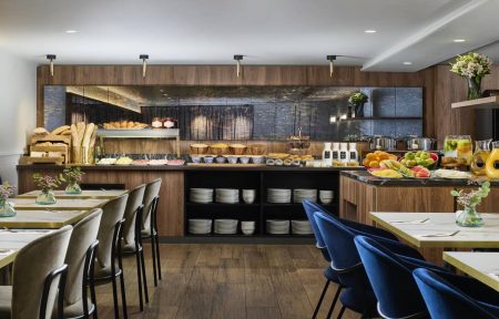 Restaurant d’hôtel pour groupes avec buffet en bois et pierre sombre proposant pains, viennoiseries, céréales, yaourts, fromages, charcuteries, fruits frais et jus, avec tables claires et assises en velours bleu.