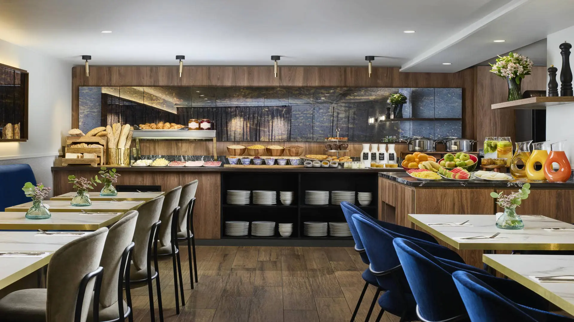 Restaurant d’hôtel pour groupes avec buffet en bois et pierre sombre proposant pains, viennoiseries, céréales, yaourts, fromages, charcuteries, fruits frais et jus, avec tables claires et assises en velours bleu.