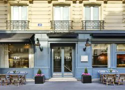 Élégante façade d’hôtel parisien aux teintes bleu clair raffinées, ornée de moulures architecturales classiques. L’entrée est soulignée par un auvent noir et des lanternes murales. L’enseigne « NH Opéra-Faubourg » est visible sur les vitres.