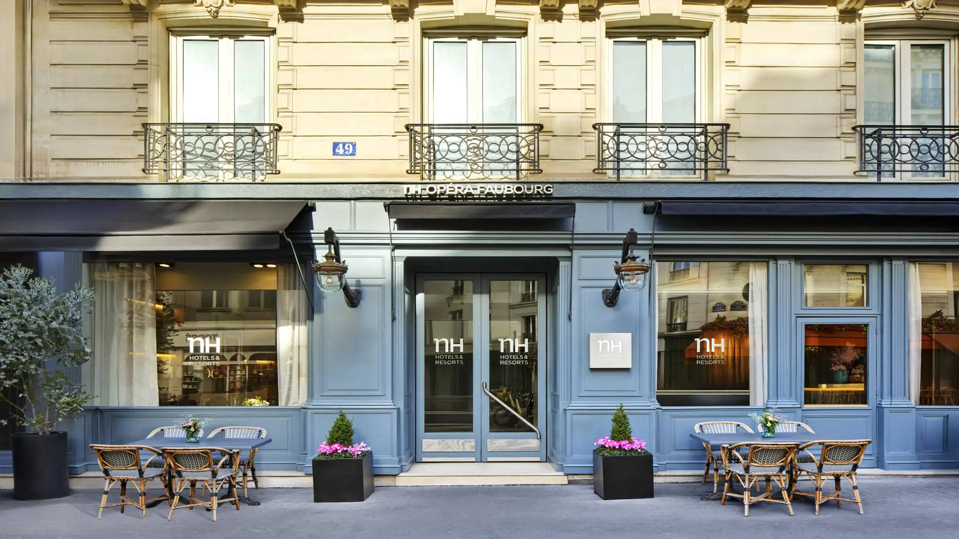 Élégante façade d’hôtel parisien aux teintes bleu clair raffinées, ornée de moulures architecturales classiques. L’entrée est soulignée par un auvent noir et des lanternes murales. L’enseigne « NH Opéra-Faubourg » est visible sur les vitres.