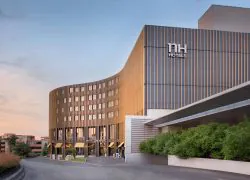 Imposante façade de l’hôtel NH Toulouse Airport, habillée d’un revêtement à l’effet lames de bois. Elle présente plusieurs fenêtres correspondant aux chambres, ainsi que de grandes baies vitrées donnant sur le restaurant. Un parvis longe le bâtiment et se prolonge vers un second espace abritant les salles de réunion, également doté de larges baies vitrées. Une rangée de petits arbustes fleuris borde ce passage, formant un chemin végétalisé qui délimite l’espace et préserve l’intimité avant l’entrée du second bâtiment.