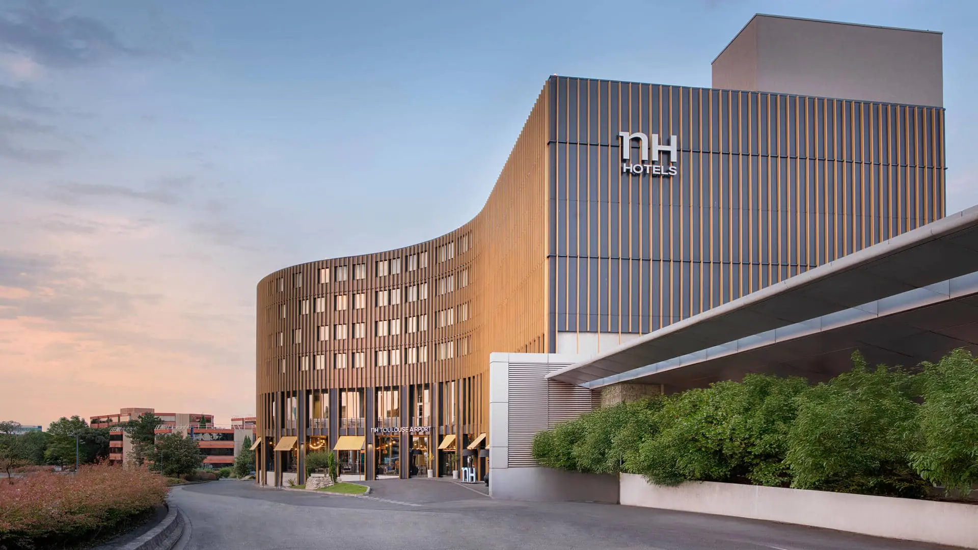 Imposante façade de l’hôtel NH Toulouse Airport, habillée d’un revêtement à l’effet lames de bois. Elle présente plusieurs fenêtres correspondant aux chambres, ainsi que de grandes baies vitrées donnant sur le restaurant. Un parvis longe le bâtiment et se prolonge vers un second espace abritant les salles de réunion, également doté de larges baies vitrées. Une rangée de petits arbustes fleuris borde ce passage, formant un chemin végétalisé qui délimite l’espace et préserve l’intimité avant l’entrée du second bâtiment.