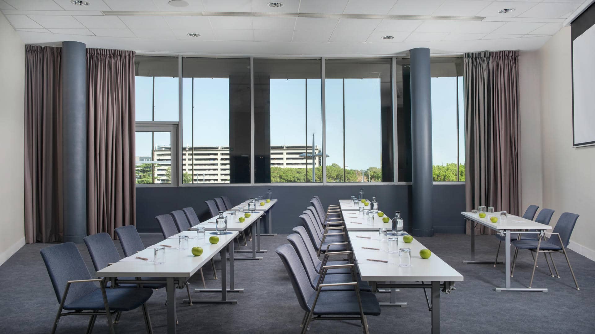 Salle de réunion aménagée en configuration conférence, avec plusieurs longues tables rectangulaires blanches disposées en rangées parallèles et entourées de chaises en tissu gris bleuté. Chaque place est équipée d’un verre, d’une bouteille d’eau, d’un stylo et d’une pomme verte. Au fond, de grandes baies vitrées offrent une vue sur des bâtiments et des arbres. Des rideaux gris taupe encadrent les fenêtres, tandis que deux colonnes cylindriques gris foncé se détachent devant celles-ci. Le sol est recouvert d’une moquette gris ardoise, les murs sont blancs et le plafond à dalles blanches intègre des spots lumineux. Un écran de projection est visible sur le mur à droite.