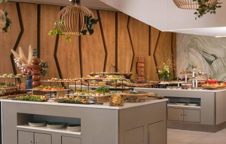 Espace buffet d’hôtel composé de plusieurs comptoirs rectangulaires beige taupe, aux plateaux blancs, présentant une grande variété de préparations salées, de salades, de verrines, de pièces apéritives et de desserts. Des présentoirs à plusieurs niveaux permettent de disposer les aliments en hauteur. Des assiettes sont rangées sur les étagères intégrées aux comptoirs. À l’arrière, le mur est habillé de panneaux en bois brun clair, structurés par des lignes géométriques noires. À droite, un mur décoratif effet pierre présente des motifs ondulés dans des tons gris, beige et blanc. Des suspensions lumineuses en forme de cages arrondies, aux finitions bois et métal noir, sont agrémentées de plantes vertes retombantes. Des compositions florales et végétales décorent les buffets. Le sol est recouvert de grands carreaux beige clair.