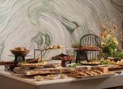 Buffet de restauration de l’hôtel composé de plusieurs plateaux présentant un assortiment de charcuteries, de fromages, de salades et de préparations froides. Les mets sont disposés sur des supports à différents niveaux, mêlant plateaux noirs, présentoirs métalliques et planches en bois naturel. À l’arrière, un grand mur décoratif aux motifs ondulés mêle des nuances de beige, de gris, de vert sauge et de mauve. Sur la droite, une composition florale aux tons jaunes, orangés et verts apporte une touche colorée. L’ensemble repose sur un comptoir aux façades beige taupe et au plateau blanc.